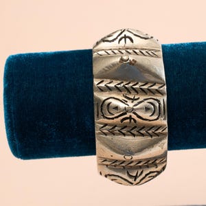 Könnte beinhalten: Ein silbernes Armband mit komplizierten geometrischen Mustern. Das Armband ist breit und hat eine abgerundete Form. Es wird auf einem blauen Samtständer präsentiert.