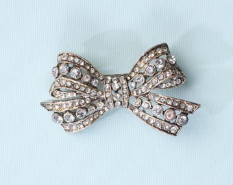 Puede incluir: Broche plateado con forma de lazo, incrustado con numerosos brillantes redondos y transparentes. El broche está sobre un fondo azul claro. El diseño del lazo presenta detalles metálicos y un estilo clásico y elegante.