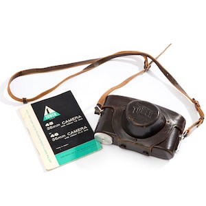 May include: A brown leather camera case with a black camera inside. The camera has a black leather strap and a silver lens. The case has a silver buckle and a silver strap. The camera is labeled "Tower 45 35mm Camera with  vector f/2 lens".
