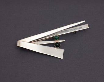 May include: A silver-toned brooch with an abstract design. The brooch features geometric shapes and two small, round gemstones, one green and one black. The brooch has a pin closure on the back.