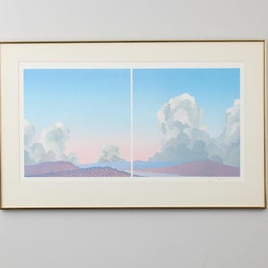 May include: A framed print of a landscape with a blue sky and white clouds. The clouds are layered and appear to be moving across the sky. The landscape is a muted purple and pink color.