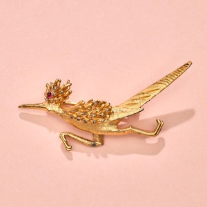 May include: A gold-toned brooch in the shape of a roadrunner with a red jewel for an eye.