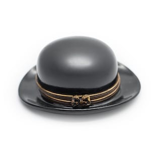 May include: A small, black, rounded hat with a smooth, glossy finish. The hat features a decorative band made of thin, metallic bronze-colored wire wrapped around the base, accented with a small, centered bow detail in the same metallic tone.