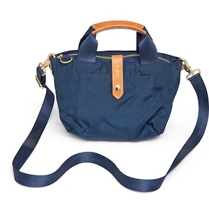 May include: A navy blue nylon handbag featuring two short top handles with tan leather accents, a detachable navy blue shoulder strap, gold-tone hardware, and a central vertical tan leather tab with a gold snap closure and the embossed text NIS.