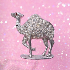 May include: Silver-toned camel brooch, encrusted with clear rhinestones. The camel has a red rhinestone eye and is standing on a small, oval base. A vintage-style accessory.