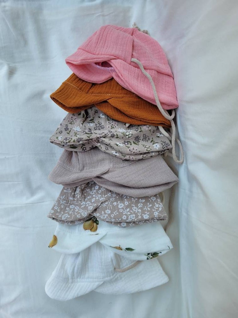 May include: A stack of five different sun hats. The top hat is pink, the second is brown, the third is beige with a floral pattern, the fourth is beige, and the bottom hat is white with a lemon pattern.