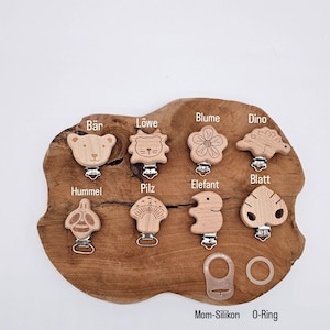 May include: Wooden teether clips with various animal and nature designs, including a bear, lion, flower, dinosaur, bee, mushroom, elephant, and leaf. The clips are displayed on a wooden surface with the text "Mom-Silikon O-Ring Adapter f&uuml;r Knopfschnuller".