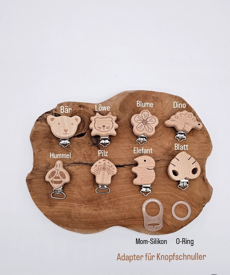 May include: Wooden teether clips with various animal and nature designs, including a bear, lion, flower, dinosaur, bee, mushroom, elephant, and leaf. The clips are displayed on a wooden surface with the text "Mom-Silikon O-Ring Adapter f&uuml;r Knopfschnuller".