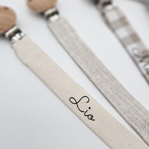 May include: Three wooden bead pacifier clips with fabric straps. One strap is white with the name "Lio" written on it, one is a beige and white checkered pattern, and one is a solid beige.