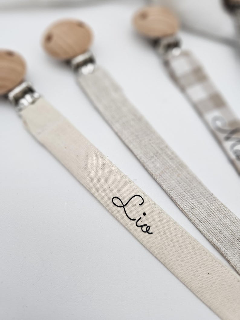 May include: Three wooden bead pacifier clips with fabric straps. One strap is white with the name "Lio" written on it, one is a beige and white checkered pattern, and one is a solid beige.