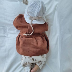 May include: A baby wearing a brown linen dress with a white crocheted hat and white pants with a brown eucalyptus leaf pattern.