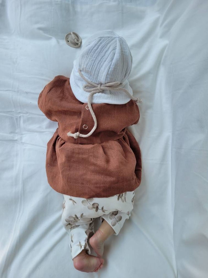 May include: A baby wearing a brown linen dress with a white crocheted hat and white pants with a brown eucalyptus leaf pattern.
