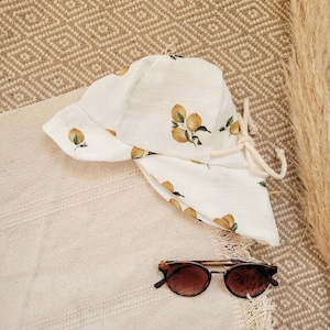 May include: A white sun hat with a lemon print and a white drawstring. The hat is lying on a beige woven rug with brown sunglasses next to it.
