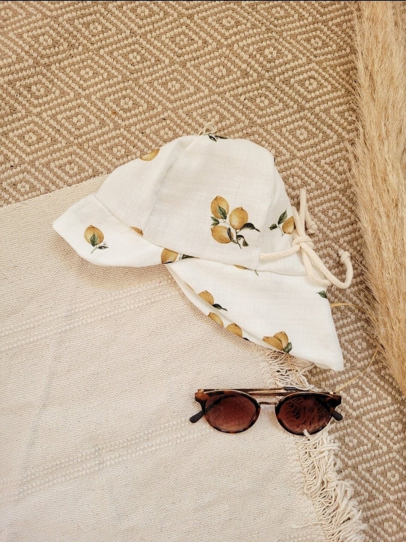 May include: A white sun hat with a lemon print and a white drawstring. The hat is lying on a beige woven rug with brown sunglasses next to it.