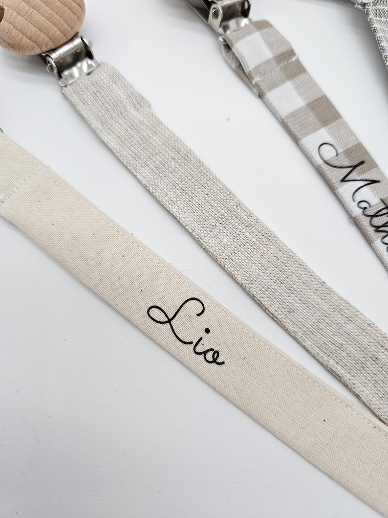 May include: Three personalized pacifier clips with wooden beads and different fabric patterns. The clips are labeled "Lio", "Mathew", and "Noah".