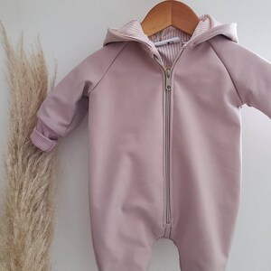 以下が含まれることがあります： 薄いピンクのベビーロンパース。ジッパーとフード付き。ロンパースはリブ編みで、木製ハンガーにかけられています。