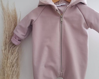 Peut inclure: Une grenouillère bébé rose pâle avec une fermeture éclair et une capuche. La grenouillère a une texture côtelée et est suspendue à un cintre en bois.