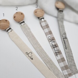 May include: Four personalized pacifier clips with wooden beads and different fabric ribbons. The ribbons are beige, silver, beige and white gingham, and gray. The names "Lio", "Mathilda", "Thomas", and the number "4" are written on the ribbons.