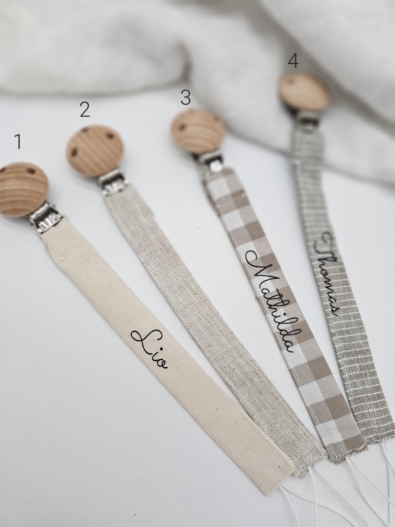 May include: Four personalized pacifier clips with wooden beads and different fabric ribbons. The ribbons are beige, silver, beige and white gingham, and gray. The names "Lio", "Mathilda", "Thomas", and the number "4" are written on the ribbons.