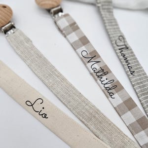 May include: Three wooden teethers with fabric straps. The straps are beige and white with the names "Lio", "Mathilda", and "Thomas" written on them.