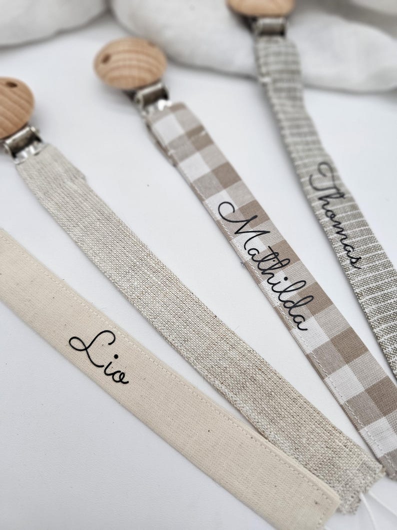 May include: Three wooden teethers with fabric straps. The straps are beige and white with the names "Lio", "Mathilda", and "Thomas" written on them.