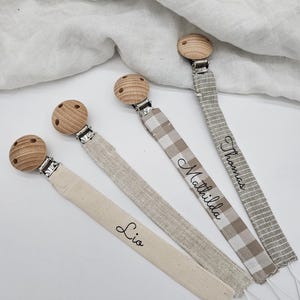 May include: Three wooden baby teethers with clips and personalized fabric straps. The straps are in different patterns and colors, including beige, white, and brown. The names "Lio", "Mathilda", and "Thomas" are embroidered on the straps.