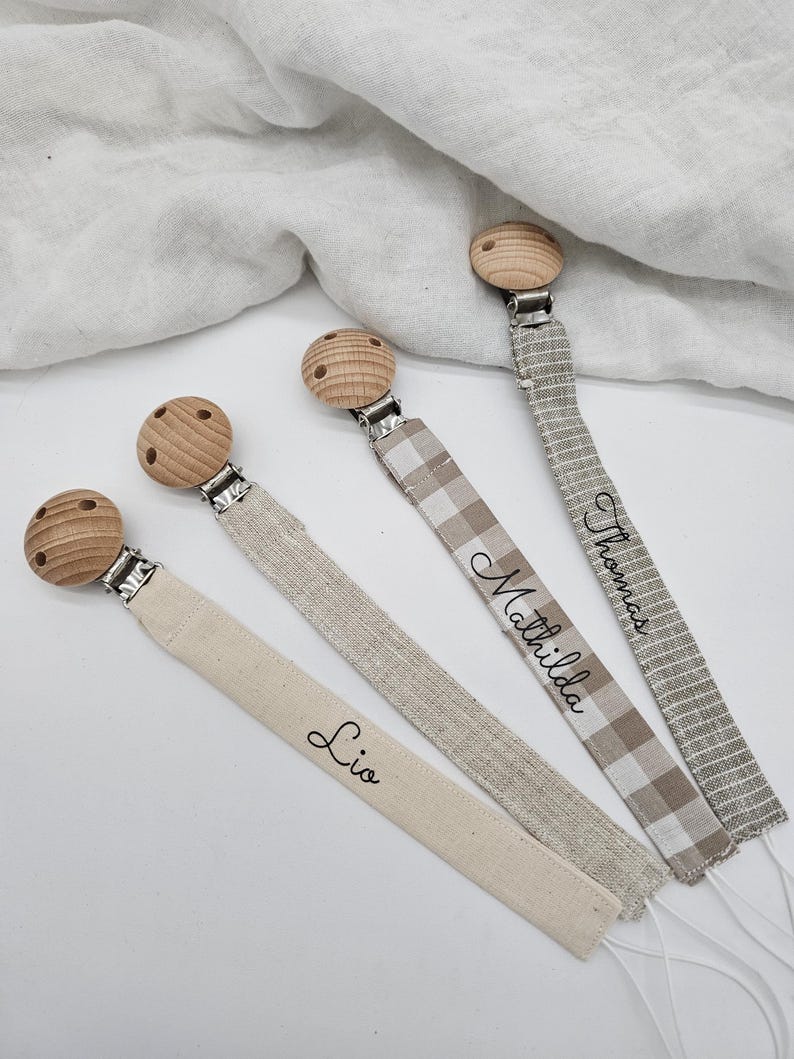 May include: Three wooden baby teethers with clips and personalized fabric straps. The straps are in different patterns and colors, including beige, white, and brown. The names "Lio", "Mathilda", and "Thomas" are embroidered on the straps.