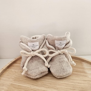 May include: A pair of light gray baby booties with cream-colored ties. Each bootie has a small white tag with the text "vintageliebe" in black. The booties are made of a soft, textured material and are displayed on a wooden tray.