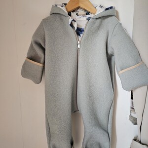 Könnte beinhalten: Hellgrauer Baby-Schneeanzug mit Kapuze und integrierten Füßen, an einem Holzbügel hängend. Der Anzug hat einen durchgehenden Reißverschluss und beige Verzierungen an den Manschetten und Füßen. Das Kapuzenfutter hat ein blau-weißes Blumenmuster.
