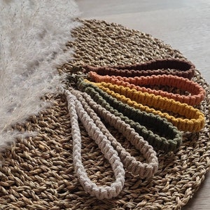 Puede incluir: Un conjunto de seis llaveros de macramé en varios tonos terrosos, incluyendo beige, marrón, verde, amarillo y naranja. Los llaveros están dispuestos en forma de abanico sobre una estera tejida.
