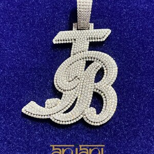 Op de afbeelding: Een met diamanten bezette hanger in de vorm van de letters "JB" op een blauwe achtergrond. De hanger heeft een oogje voor een ketting. De merknaam "anjani" staat onderaan.