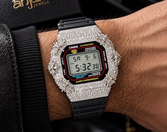 May include: A digital Casio watch with a black resin strap and a silver-tone bezel encrusted with clear stones. The watch face features a digital display showing the time 5:32 and date 9-6, with four silver cross-shaped accents on the bezel.