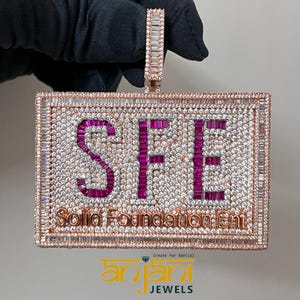 May include: A rose gold pendant with a square frame set with clear and pink baguette-shaped stones. The letters "SFE" are spelled out in pink stones inside the frame. The words "Solid Foundation Ent" are written in smaller letters below the frame.