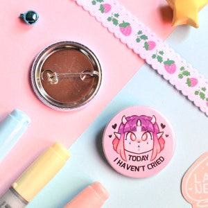 Puede incluir: Insignia de botón rosa con una ilustración de dibujos animados de un personaje de pelo rosa con cuernos y el texto "Hoy no he llorado".