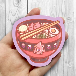 May include: A pink and red cartoon bowl of ramen with chopsticks, a boiled egg, and green onions. The bowl has a black band with purple accents and a pink cartoon creature on the front.