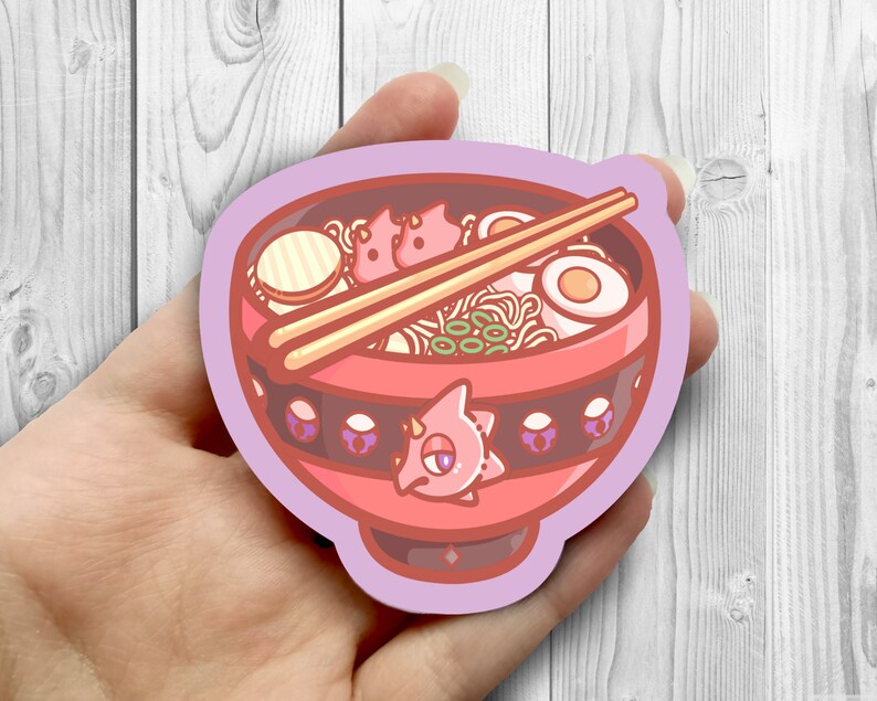 May include: A pink and red cartoon bowl of ramen with chopsticks, a boiled egg, and green onions. The bowl has a black band with purple accents and a pink cartoon creature on the front.