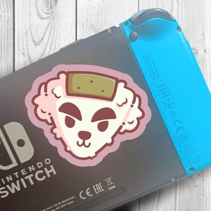 May include: A pink and white sticker of a cartoon dog with a brown patch on its head, attached to a black Nintendo Switch console with a blue Joy-Con controller.