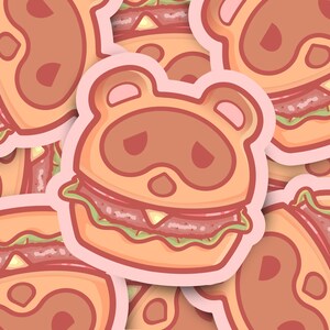May include: A sticker featuring a cartoon animal wearing a hamburger costume. The animal has brown fur, a pink nose, and black eyes. The hamburger is a classic style with a sesame seed bun, lettuce, tomato, and a beef patty.