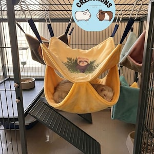 May include: A yellow ferret hammock with a ferret graphic inside a metal cage. The hammock is suspended by dark blue straps. A ferret is sleeping in the hammock. The cage also contains a ramp, food bowls, and other hammocks.
