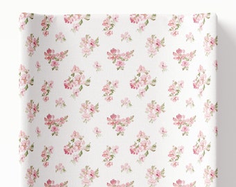 May include: A white changing pad cover with a pink floral pattern. The flowers are small and delicate, and the pattern is repeated throughout the cover.