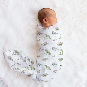 May include: A newborn baby swaddled in a white muslin swaddle blanket with a dinosaur print. The blanket features green, brown, and gray dinosaurs on a white background.