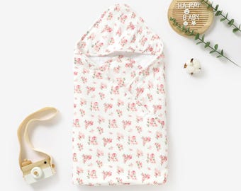 May include: A white hooded towel with a pink floral pattern. The towel is folded and laid flat on a white surface. The text "Happy Baby" is visible on a wooden sign in the background.
