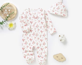 May include: A white baby onesie with a pink floral print and a matching pink floral print hat. The onesie has a zipper closure and long sleeves.