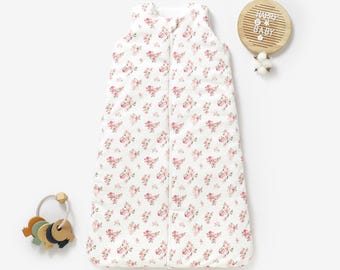 May include: A white sleeveless baby sleeping bag with a pink floral print. The sleeping bag has a zipper down the front and a white trim around the edges.