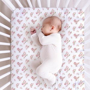 May include: A white cot with a white fitted sheet featuring a pink floral pattern. A baby is sleeping on the sheet.