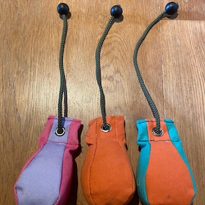 May include: Three dog training dummies in vibrant colours. One is pink and lilac, one is orange, and the third is orange and turquoise. Each has a black ball attached to a green cord.