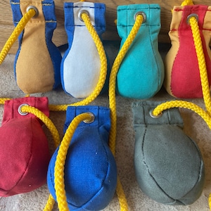 Puede incluir: Nueve bolsas de frijoles de tela de colores con cuerdas amarillas. Cada bolsa de frijoles es de un color diferente: rojo, amarillo, azul, verde, blanco, gris y dos tonos de azul.