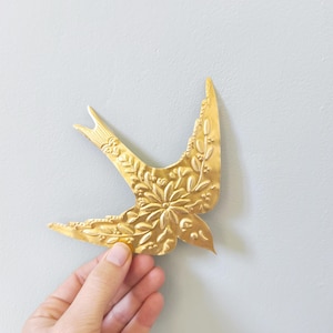 Può includere: Un ornamento a forma di uccello in metallo dorato con intricati dettagli floreali e di foglie. L'uccello è tenuto su uno sfondo grigio chiaro. Le ali dell'uccello sono spiegate e il design è in rilievo.