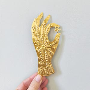 Puede incluir: Escultura de mano de metal dorado con intrincados patrones florales y de hojas. La mano sostiene un objeto en forma de estrella. La escultura es probablemente un artículo decorativo o para colgar en la pared.