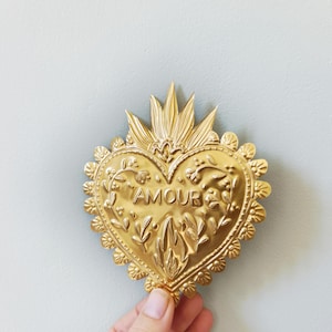 Puede incluir: Un adorno de pared dorado en forma de corazón con la palabra "AMOUR" en relieve en el centro. El corazón está adornado con diseños florales y de hojas, y un elemento en forma de corona en la parte superior. La pieza es sostenida por una mano sobre un fondo azul claro.